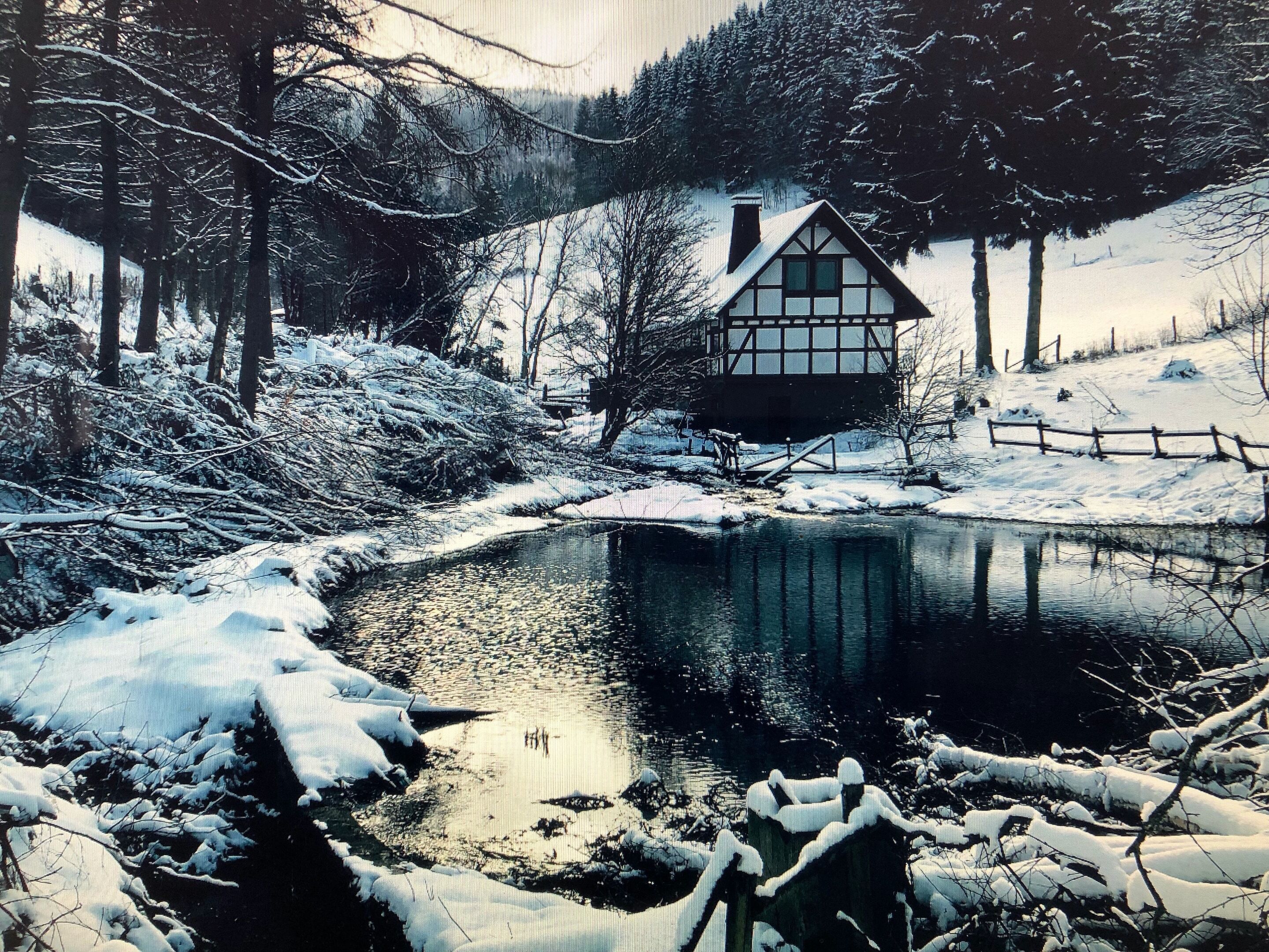 A snow-covered landscape with a small house by a reflective water body, surrounded by trees and a wooden fence.