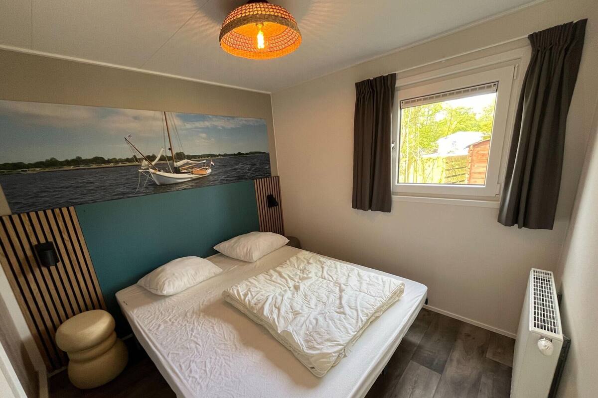 A bedroom with a bed, a painting of a boat on water, a window with curtains, and a radiator.