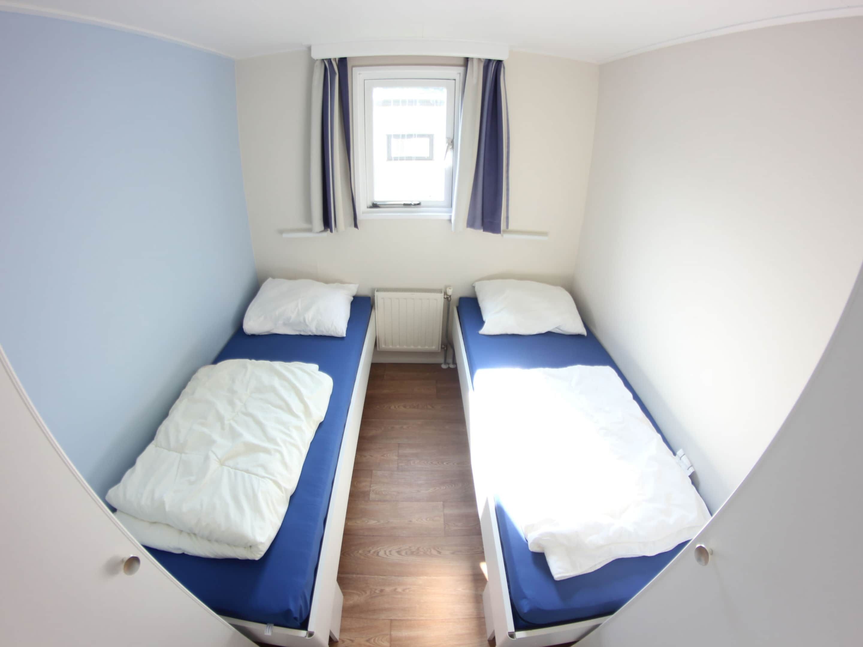 A compact room with two single beds, a window with curtains, and a radiator.