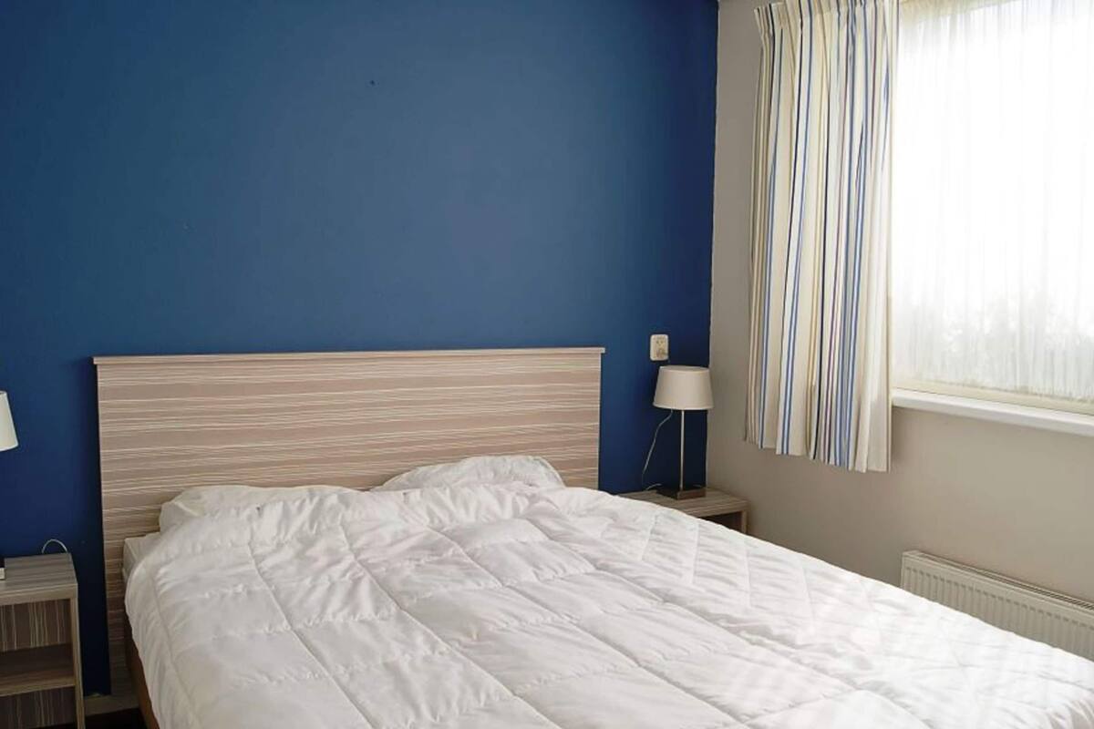 A neatly made bed in a room with a blue wall and a window with striped curtains.