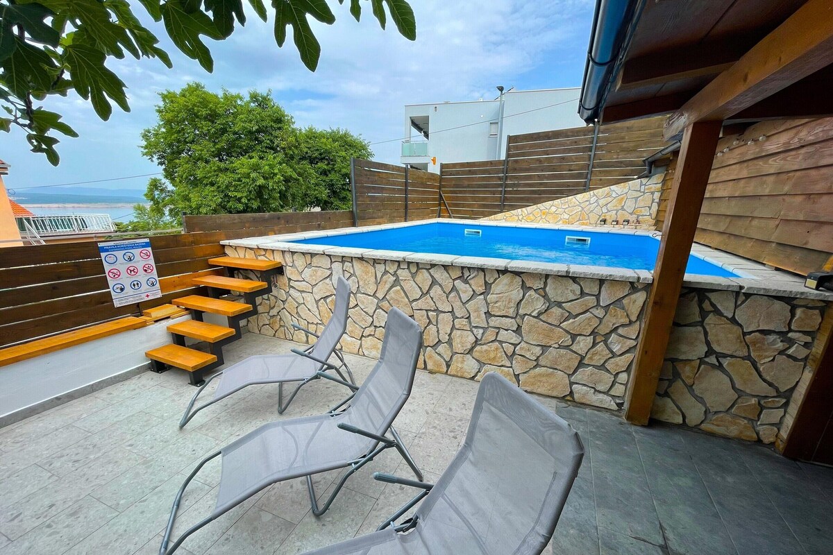 Zona de piscina con muros de piedra, terraza de madera y sillas de metal.