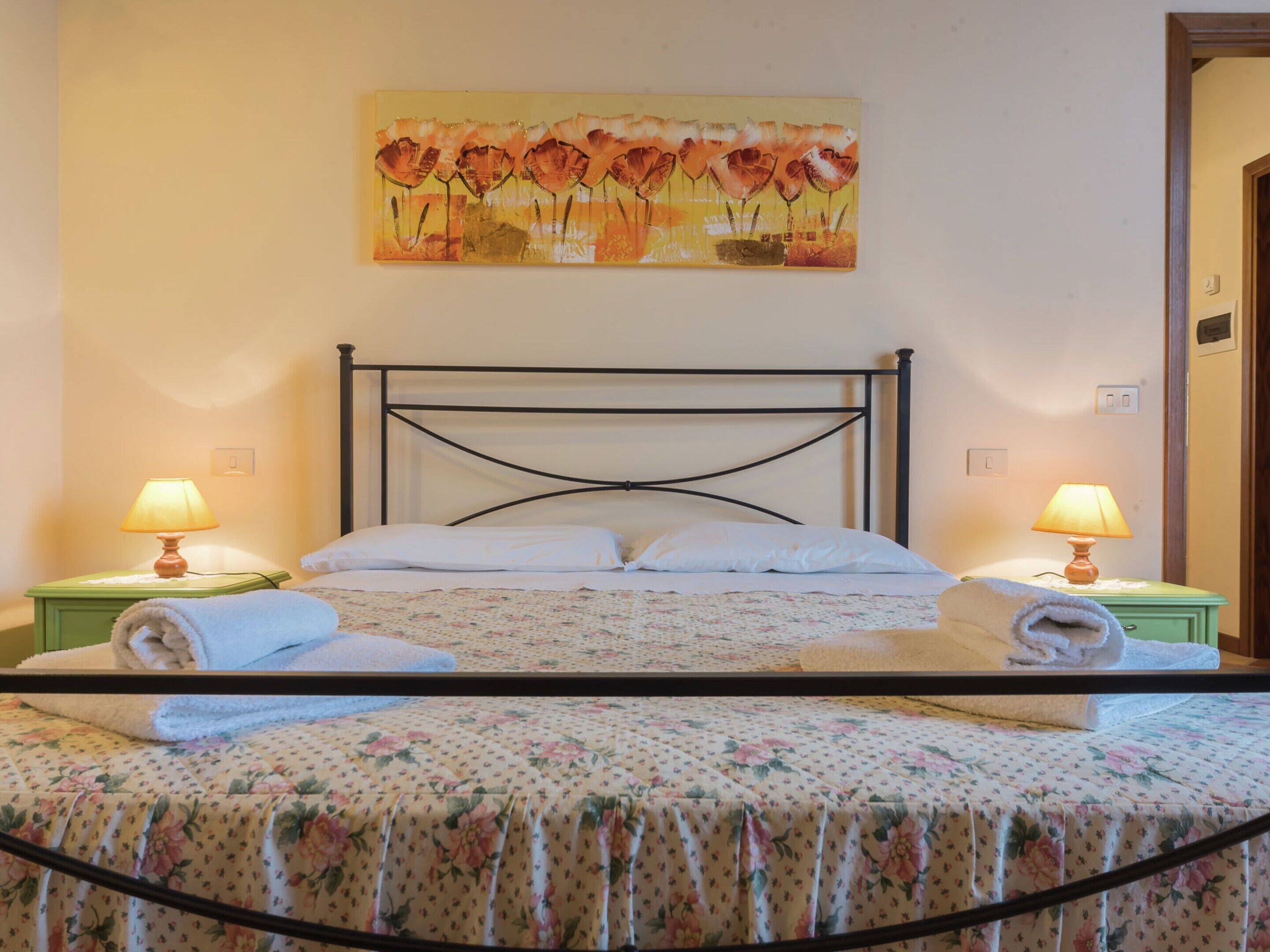 A neatly made bed with floral bedding, two bedside lamps, and a painting above the bed.