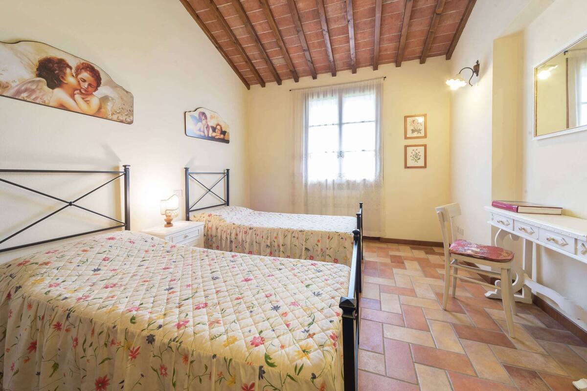 A bedroom with two beds, a wooden ceiling, a window with curtains, a bedside table, and a mirror.