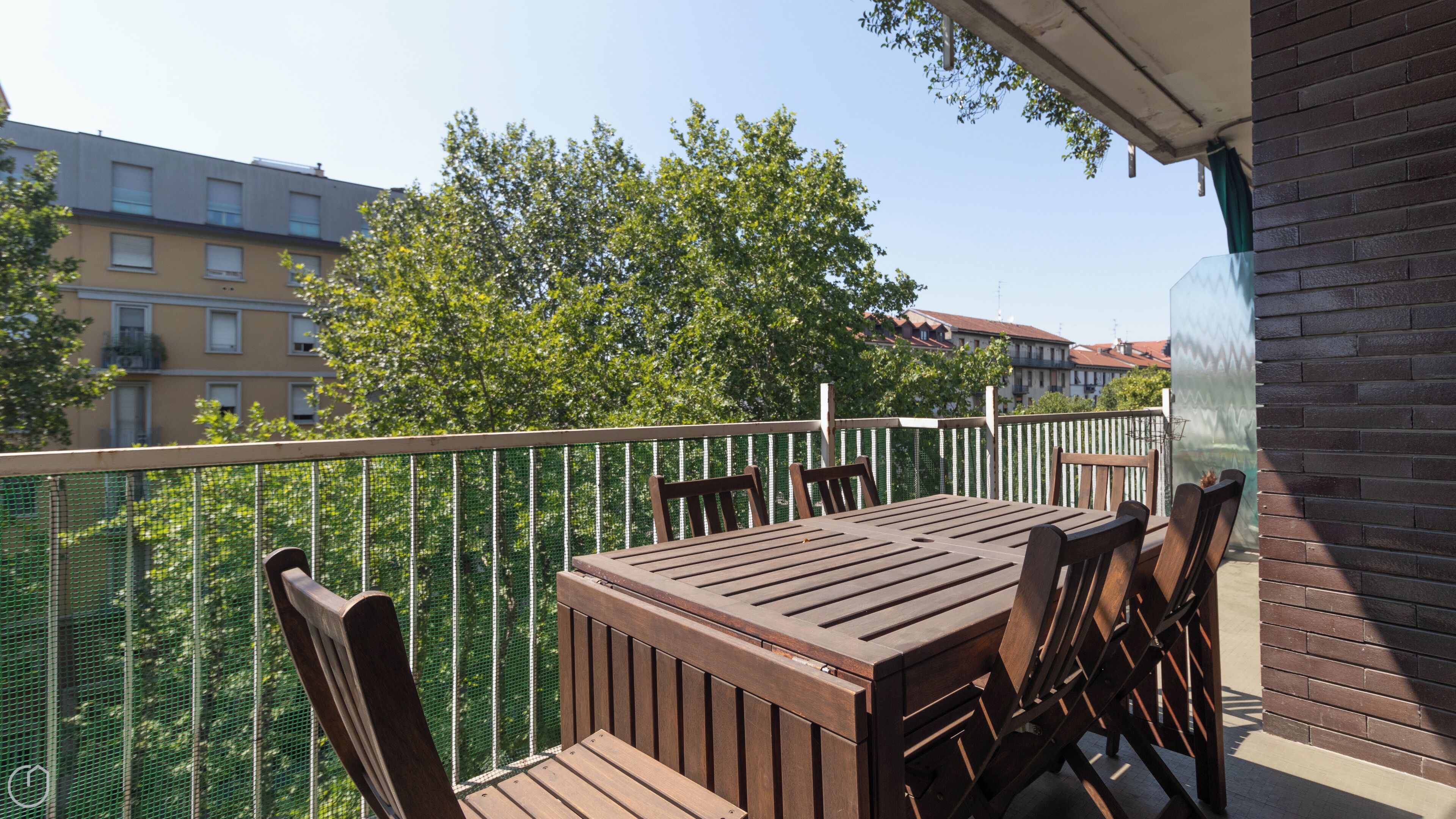 Un balcone con un tavolo e sedie di legno, una recinzione in rete verde e edifici sullo sfondo.