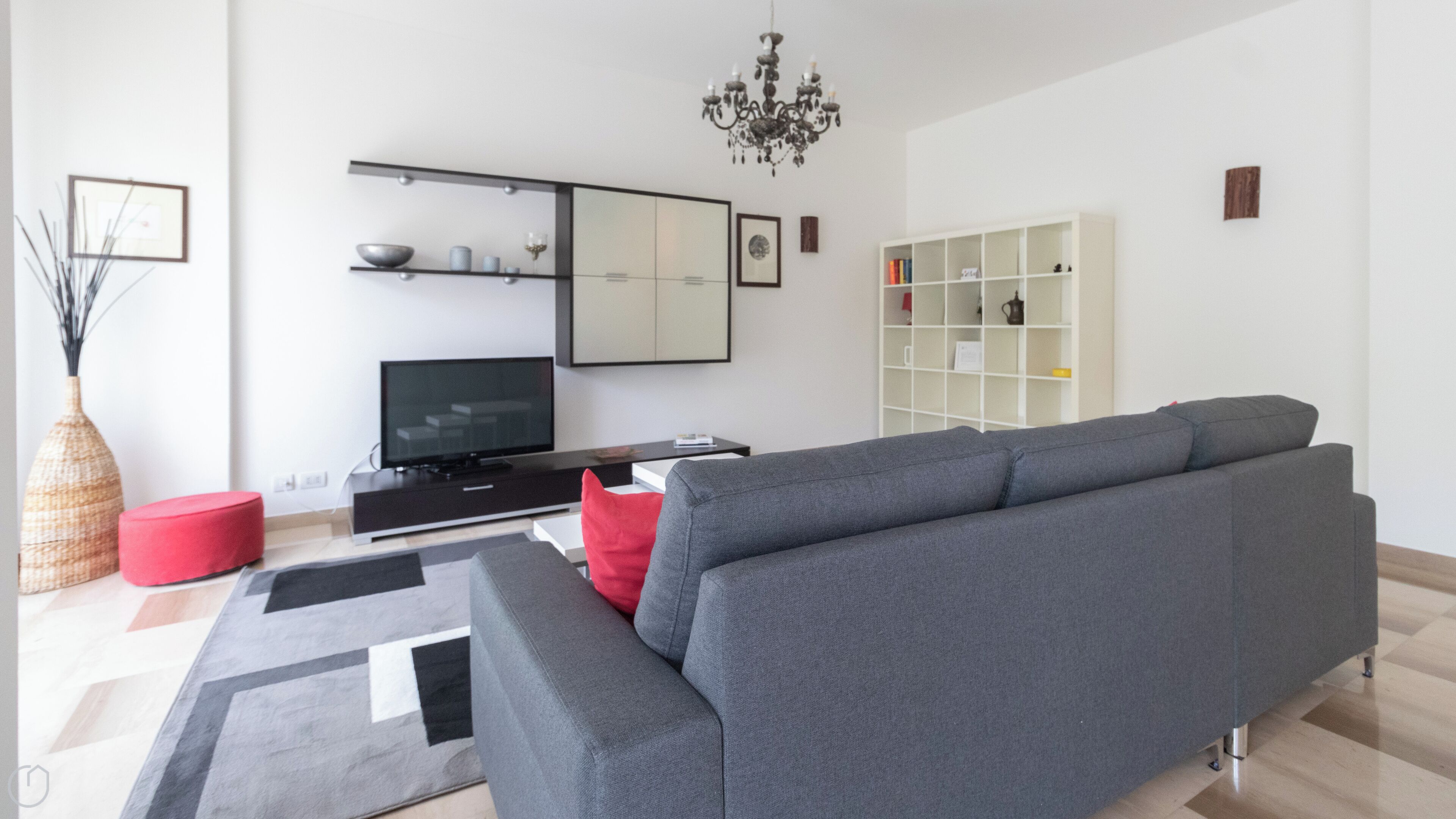 A modern living room with a grey sofa, a flat-screen TV, a red ottoman, a white shelving unit, and a chandelier.