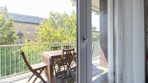 Ein Balkon mit Holztisch und -stühlen, einem Metallgeländer und Blick auf Gebäude und Bäume im Hintergrund.
