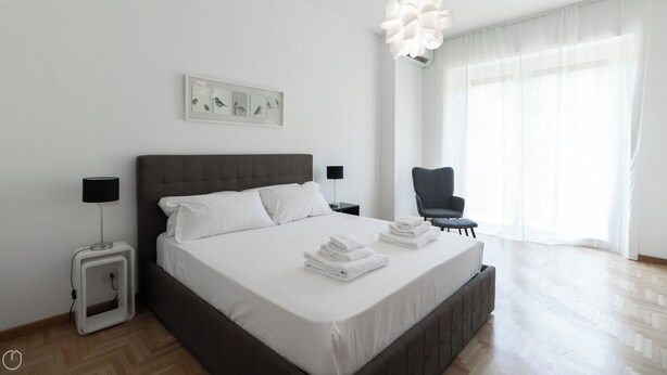Ein modernes Schlafzimmer mit einem großen Bett, einem Nachttisch mit Lampe, einem Sessel und einem Fenster mit durchsichtigen Vorhängen.