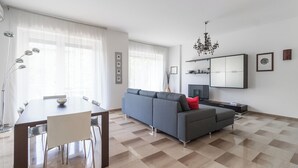Ein modernes Wohnzimmer mit einem grauen Sofa, einem Esstisch mit Stühlen, einem Fernseher und einer Kristallleuchter.