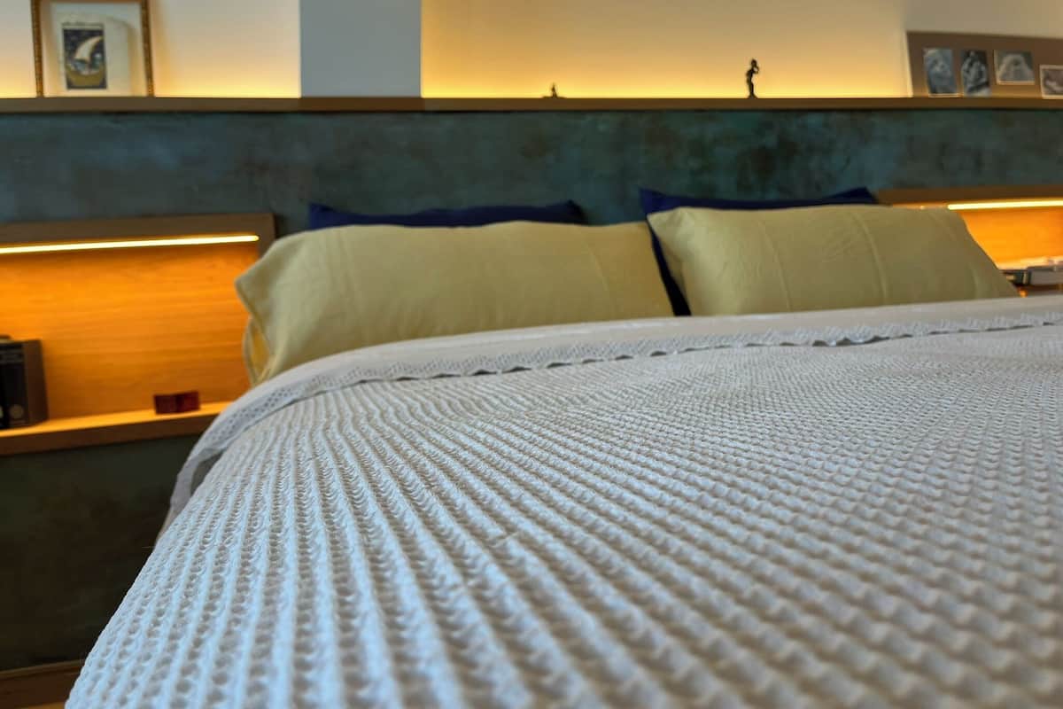 Una cama con un edredón blanco texturizado, un cabecero con diseño azul y verde, y una pared con cuadros enmarcados y un estante con un objeto pequeño.