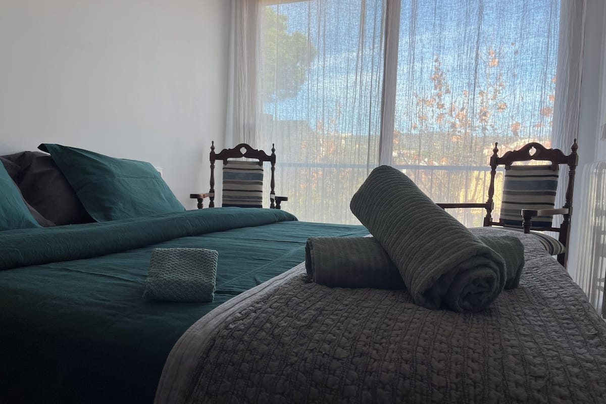 Un dormitorio con dos sillas de madera, una cama con ropa de cama verde y una ventana con cortinas de gasa.