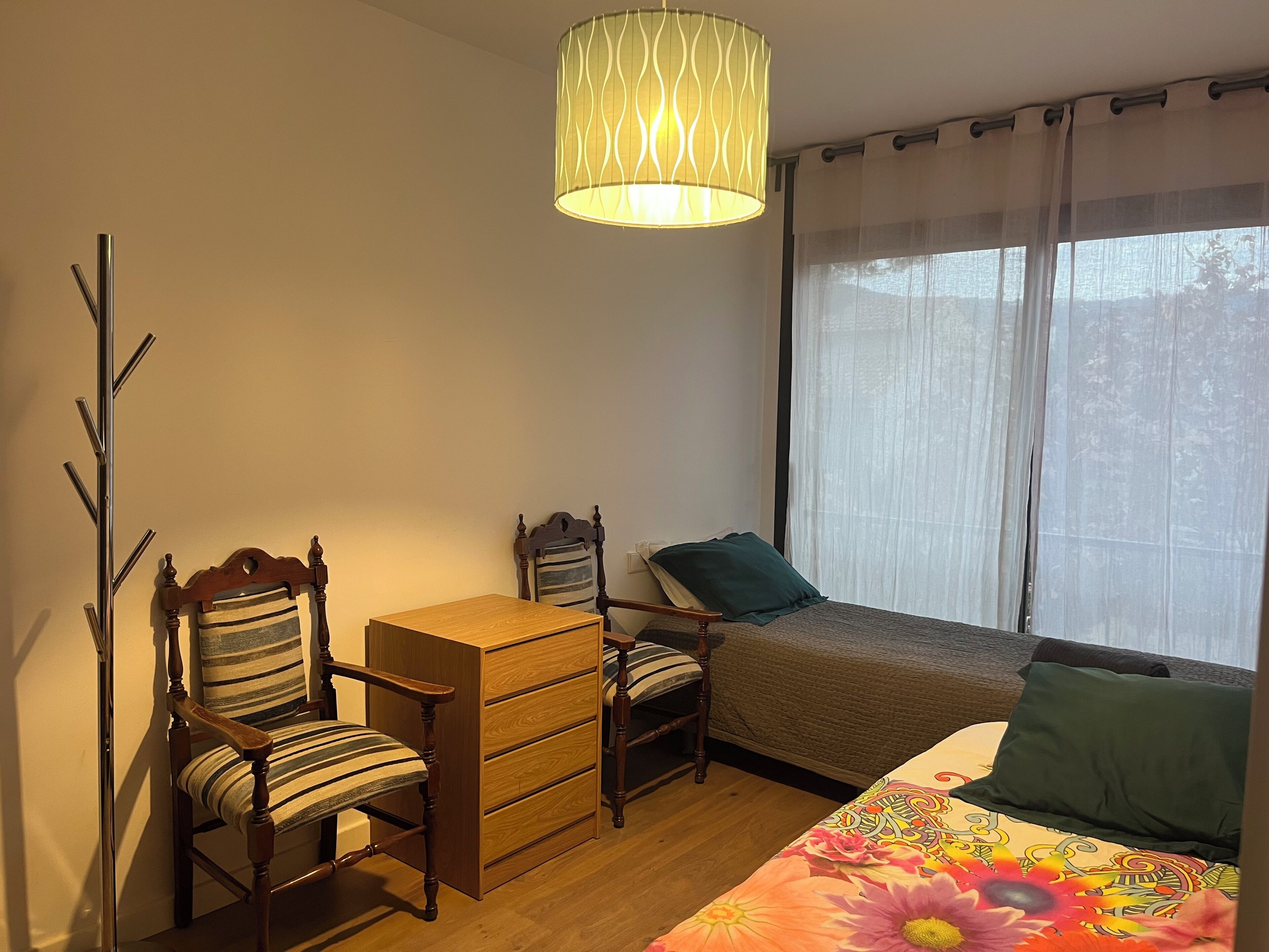 Un dormitorio con un escritorio de madera, una silla, una cama con colcha estampada y una ventana con cortinas de gasa.