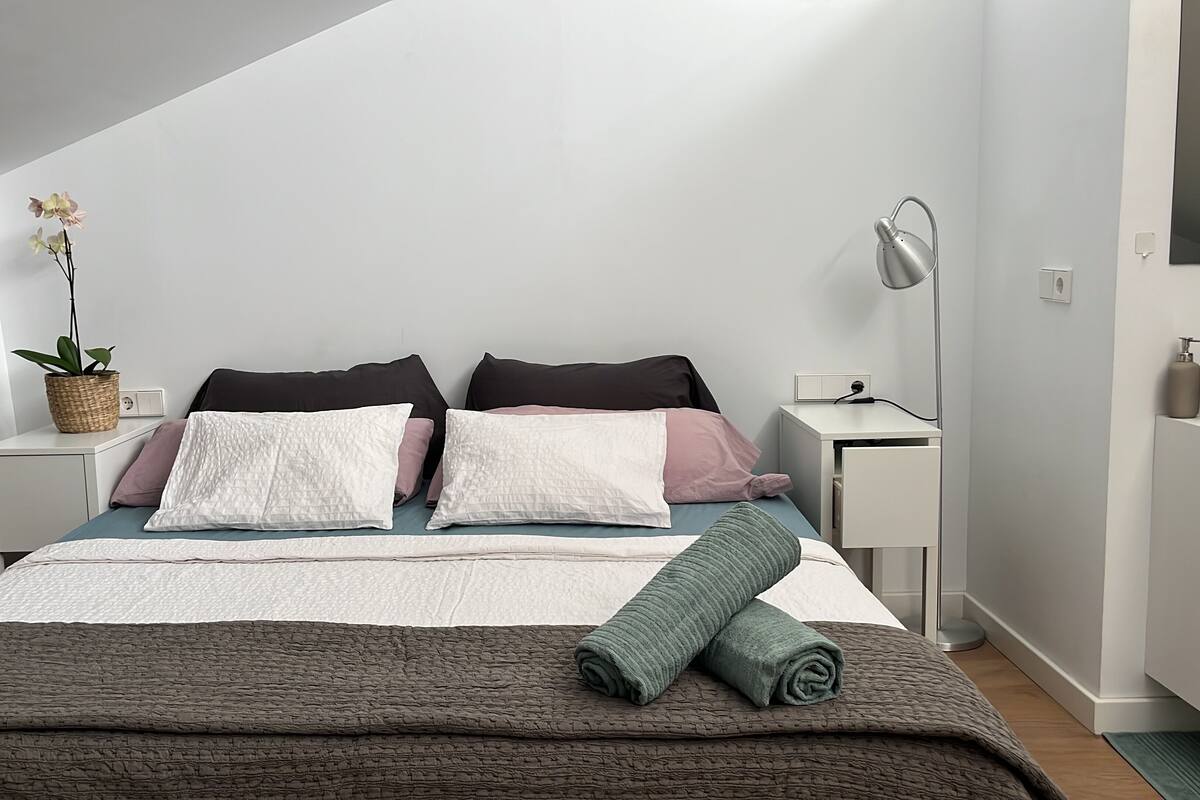 Un dormitorio ordenado con cama, almohadas, una mesita de noche, lámpara y una planta.