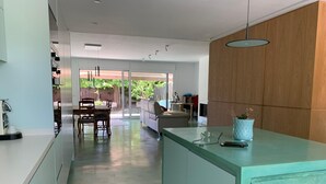 Una cocina moderna con una isla central, un comedor con mesa y sillas, y vistas a un jardín a través de puertas de cristal.