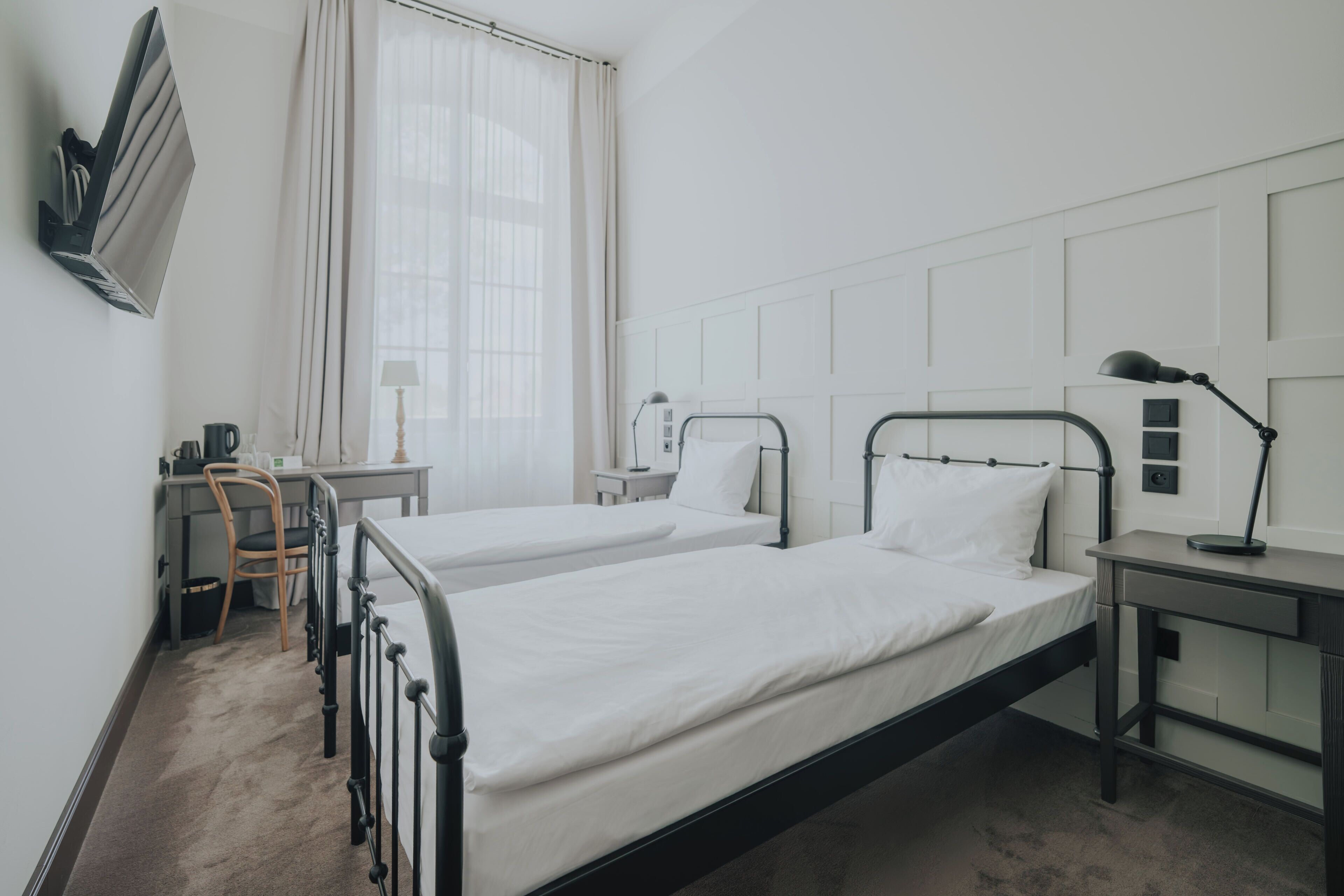 Habitación de hotel con dos camas individuales, un escritorio con silla, una lámpara y una ventana con cortinas.