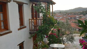 Un patio con un edificio blanco, un balcón de madera, una mesa y sillas, y una vista de un pueblo al fondo.