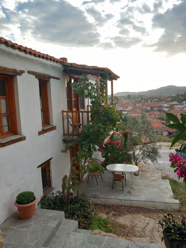 Un patio con un edificio blanco, un balcón de madera, una mesa y sillas, y una vista de un pueblo al fondo.