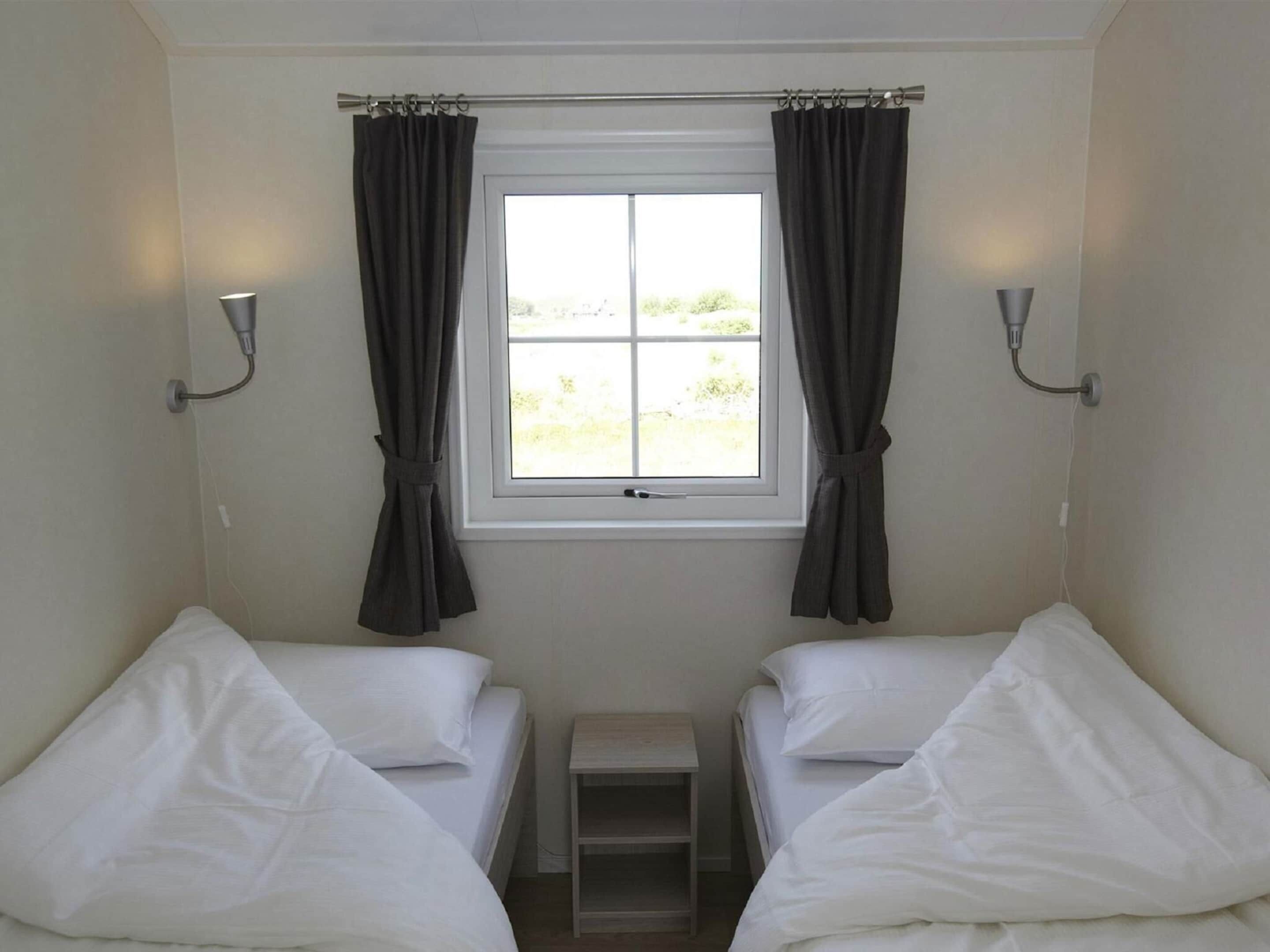 Ein kleines, sauberes Zimmer mit zwei Einzelbetten, einem Fenster mit Vorhängen und einem kleinen Tisch.