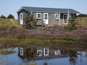 Et hus med grå facade, hvidt tagbræt og en træterrasse. Det er omgivet af lilla lyng og et vandareal, der spejler huset.