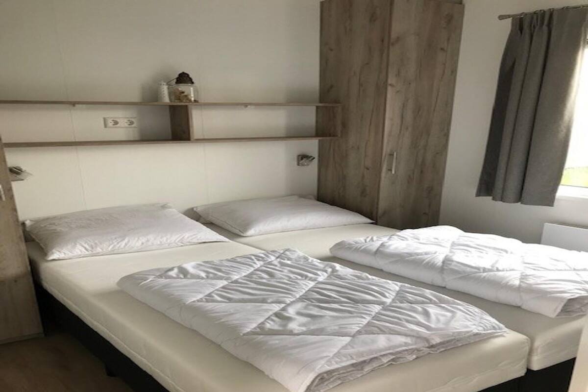 A room with two beds, a wooden wardrobe, a shelf with a decorative item, and a window with curtains.