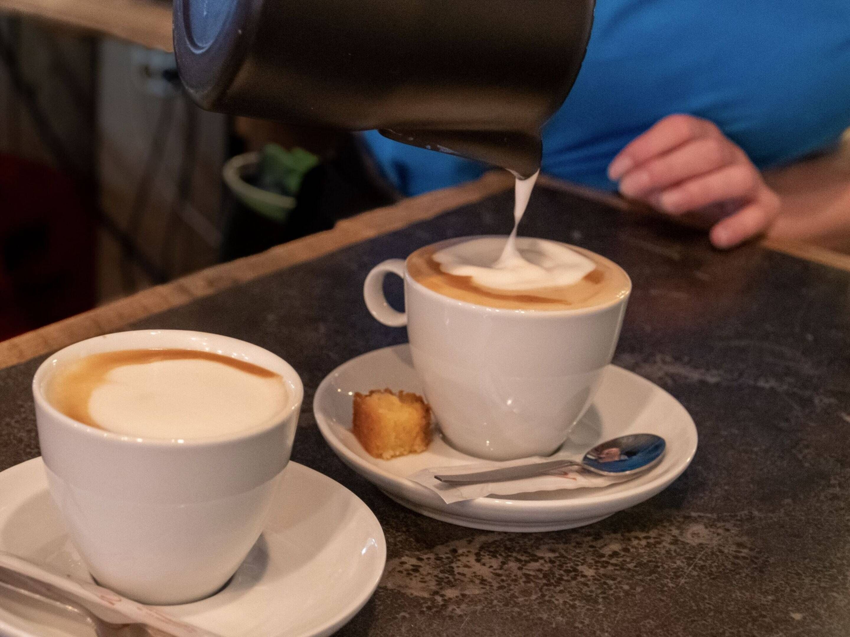 Two white cups of coffee on a table with a spoon and a small plate.
