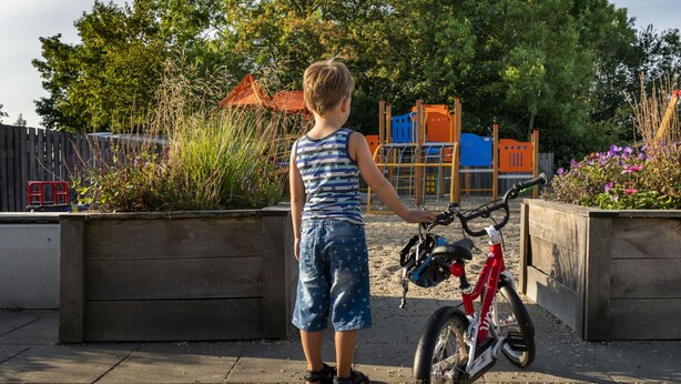 Un bambino in piedi accanto a una bicicletta rossa in un giardino con un'area giochi sullo sfondo.