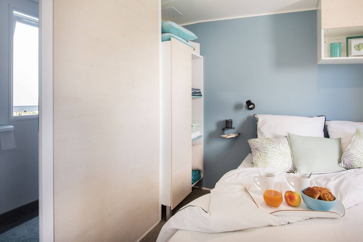 A neatly made bed with pillows, a bedside table with a lamp, and a small shelf with items.