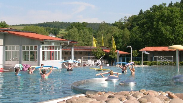 A swimming pool with people exercising, surrounded by buildings and trees. - Wooden Bungalow With a Terrace, in a Wooded Area (Ronshausen)