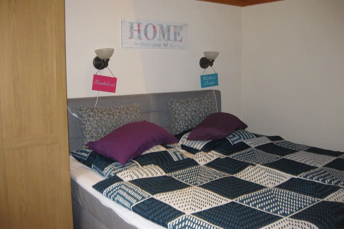 Une chambre avec un lit, deux lampes fixées au mur, une pancarte sur laquelle on peut lire « La maison, c’est là où se trouve votre cœur », et une enseigne pour le « Kuschelnest Wellness Lounge ».