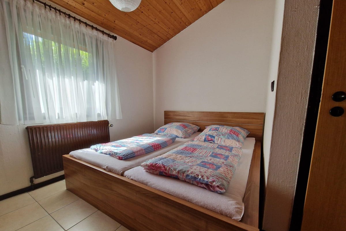 Une chambre à coucher avec un lit en bois, deux oreillers, un radiateur et une fenêtre ornée de rideaux légers.