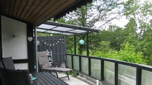 A covered patio with a table and chairs, a small bookshelf, and a decorative lantern. - Wooden Bungalow With a Terrace, in a Wooded Area (Ronshausen)