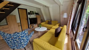 A spacious living area with a yellow sofa, a blue patterned armchair, a small round table, and a television. - Wooden Bungalow With a Terrace, in a Wooded Area (Ronshausen)