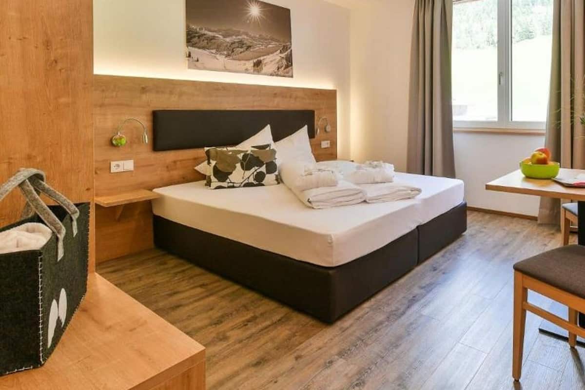 Ein modernes Hotelzimmer mit einem Holz-Kopfteil, einem großen Bett mit weißen Leinen, einem kleinen Tisch mit Obstschale und einem Fenster mit Vorhängen.