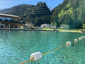 Un lac aux eaux limpides, équipé d’une corde et de bouées, entouré de bâtiments et bordé par une montagne boisée.