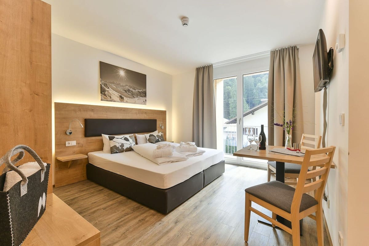 Ein modernes Hotelzimmer mit einem großen Bett, einem Holzkopfende, einem Nachttisch, einem Fernseher, einem Esstisch mit Stühlen und einem Fenster mit Vorhängen.