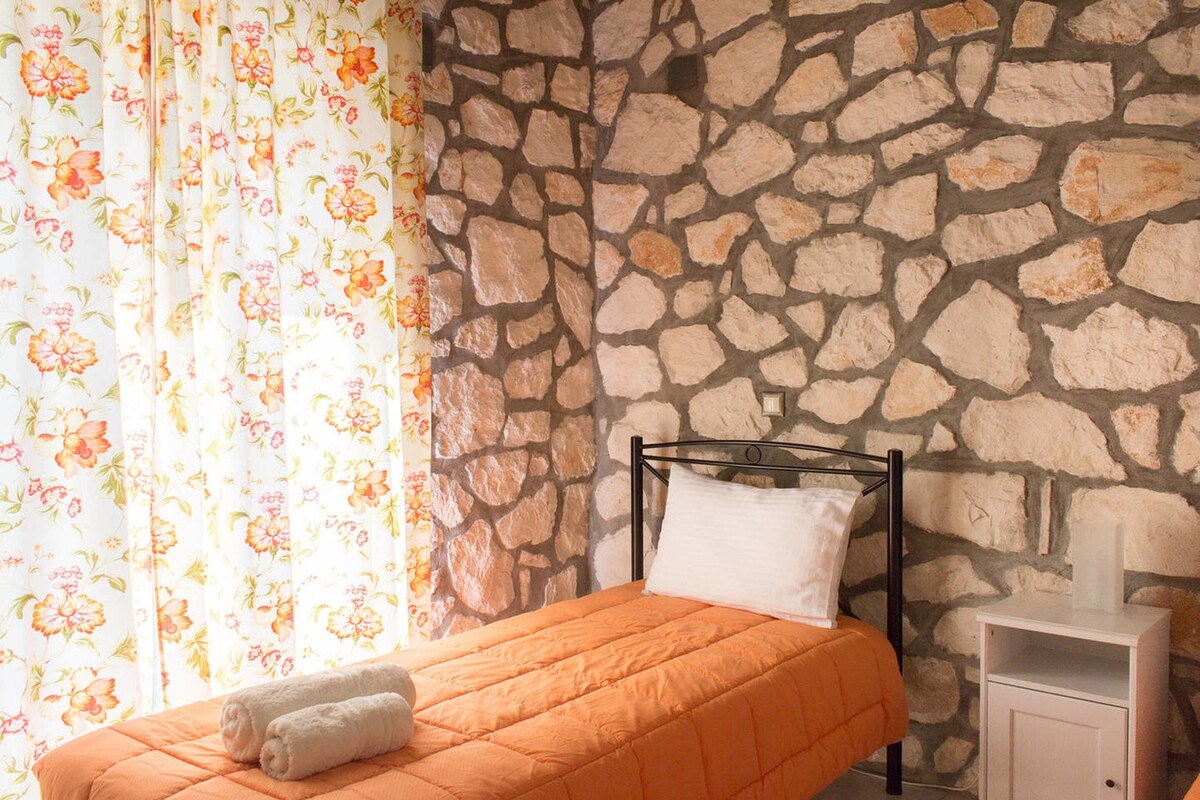 Una habitación con pared de piedra, una cama con colcha naranja, una mesita de noche con lámpara y cortinas florales.