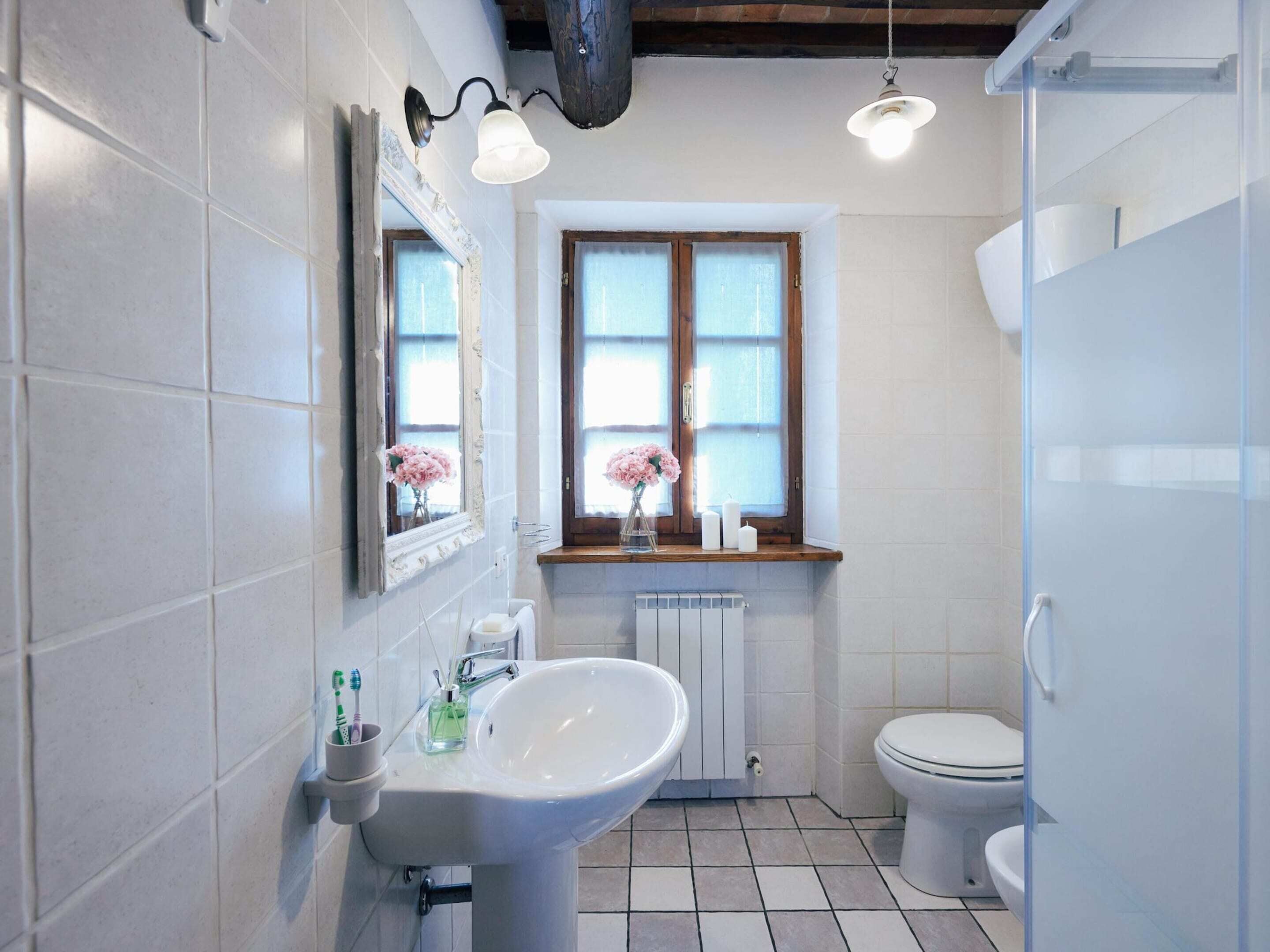 A bathroom with white tiles, a white sink, a toilet, a window with a view, and a mirror with a light fixture.