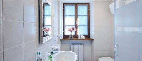A bathroom with white tiles, a white sink, a toilet, a window with a view, and a mirror with a light fixture.