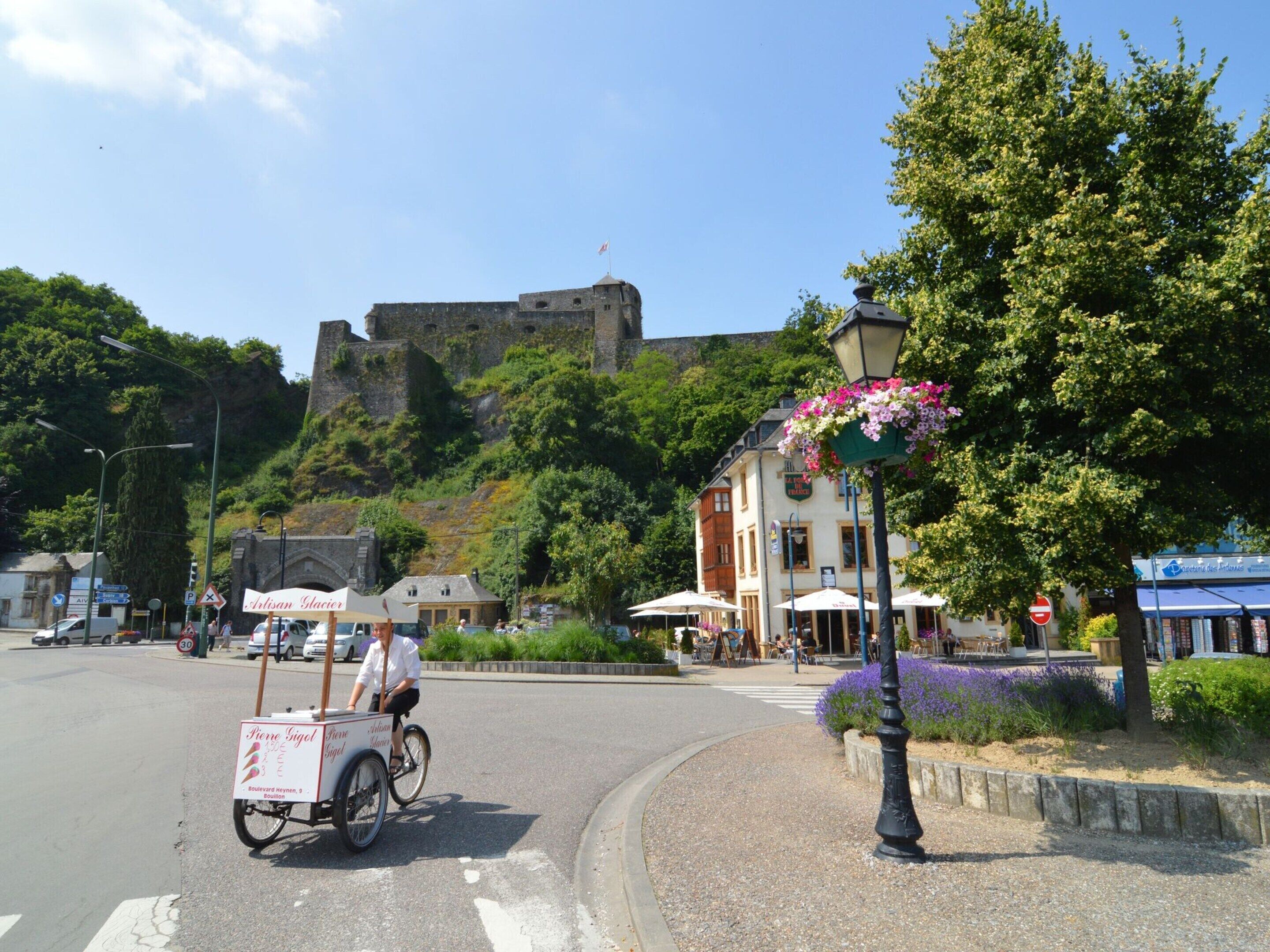 A street with a historic castle in the background, a street vendor on a bicycle, and a building with outdoor seating.