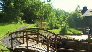 A wooden bridge over a stream with a stone path on either side, surrounded by greenery and a building in the background. - Tranquil Apartment in Sensenruth With Barbecue (Bouillon)
