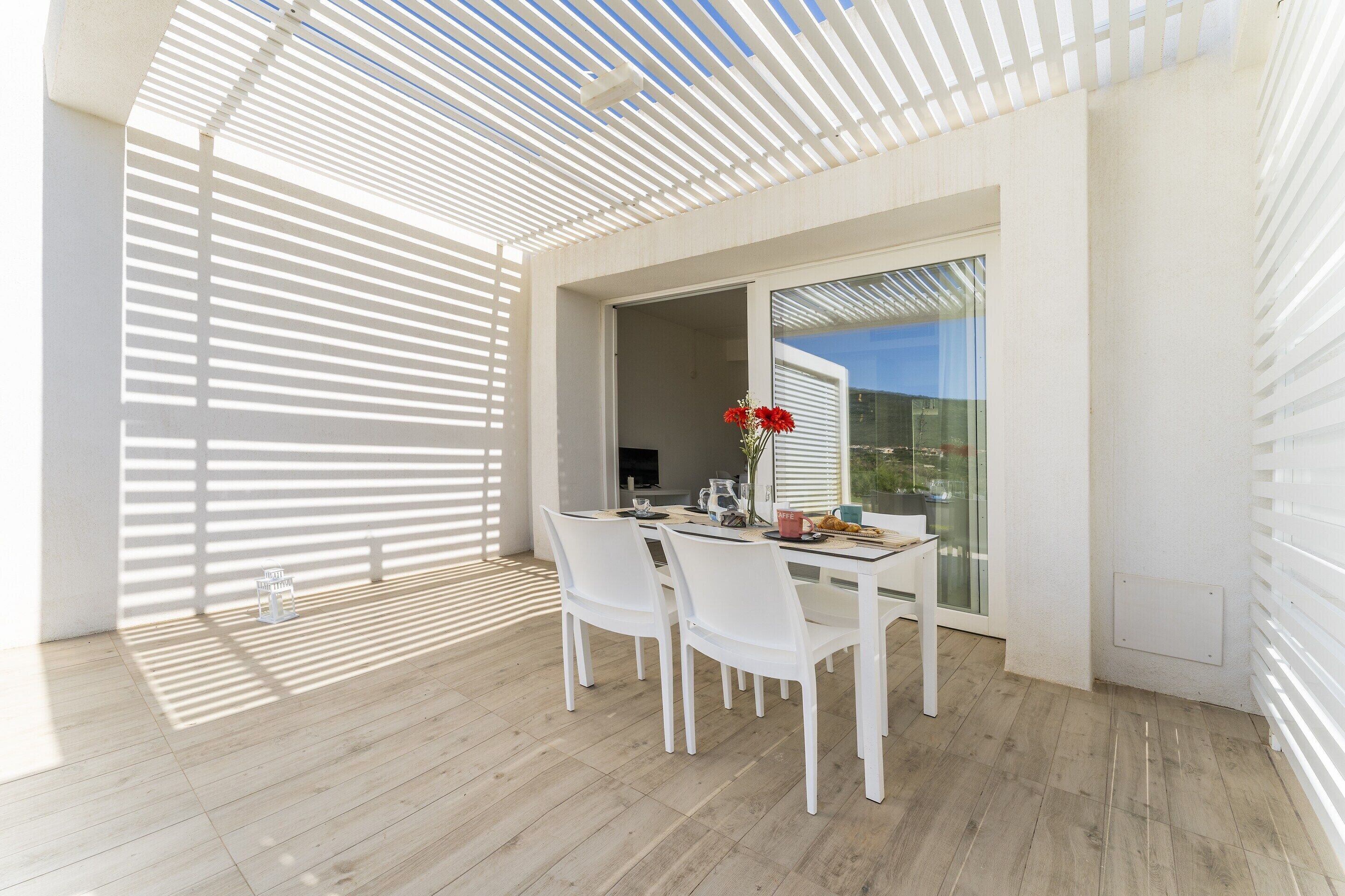A modern outdoor dining area with a white table and chairs, a glass door leading outside, and a wooden floor.