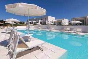 Un área de piscina con sillones de descanso y sombrillas, una piscina azul cristalina y edificios modernos al fondo.