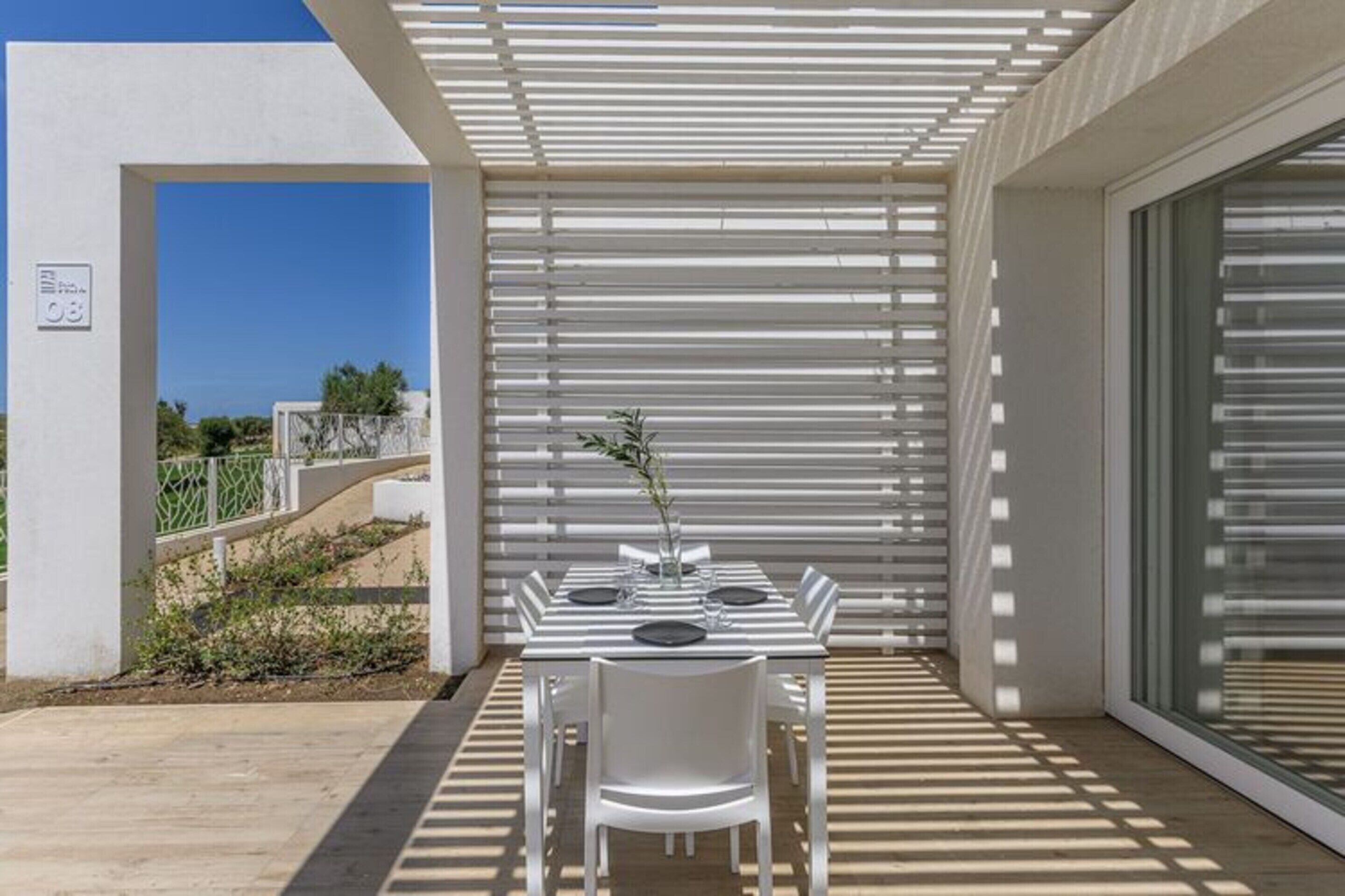 Un espace repas extérieur moderne, avec une table et des chaises blanches, une plante en pot et une vue sur la mer.
