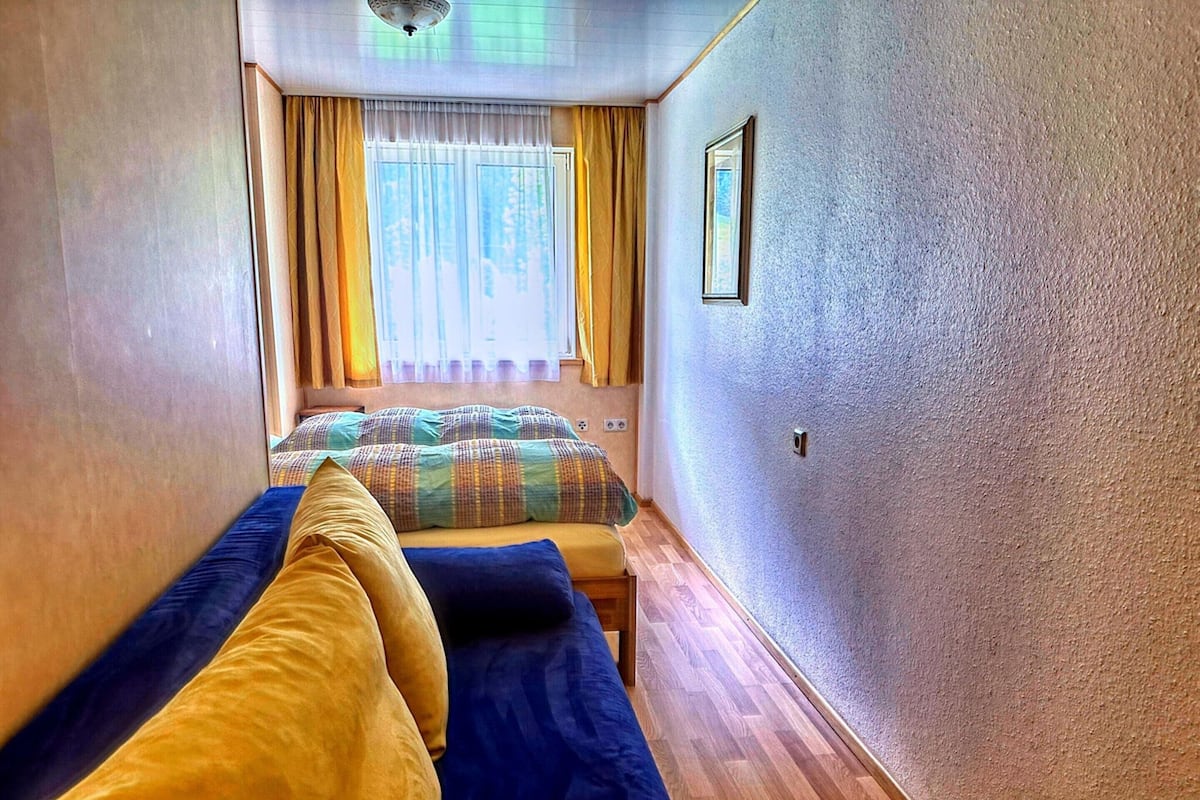 Une chambre d’hôtel étroite, avec un lit, une banquette et une fenêtre avec des rideaux.