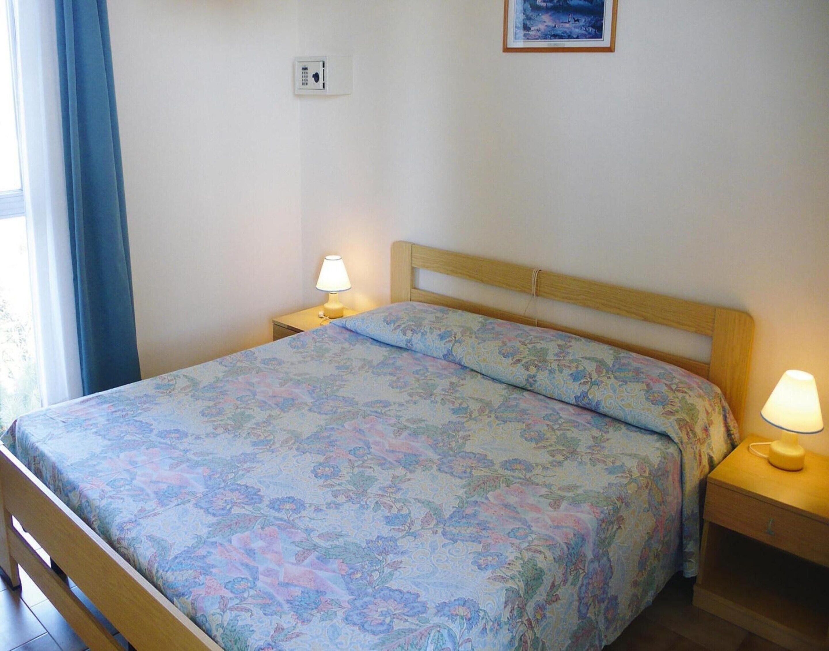 A bedroom with a floral bedspread, a wooden headboard, two bedside lamps, a nightstand, and a wall-mounted light switch.