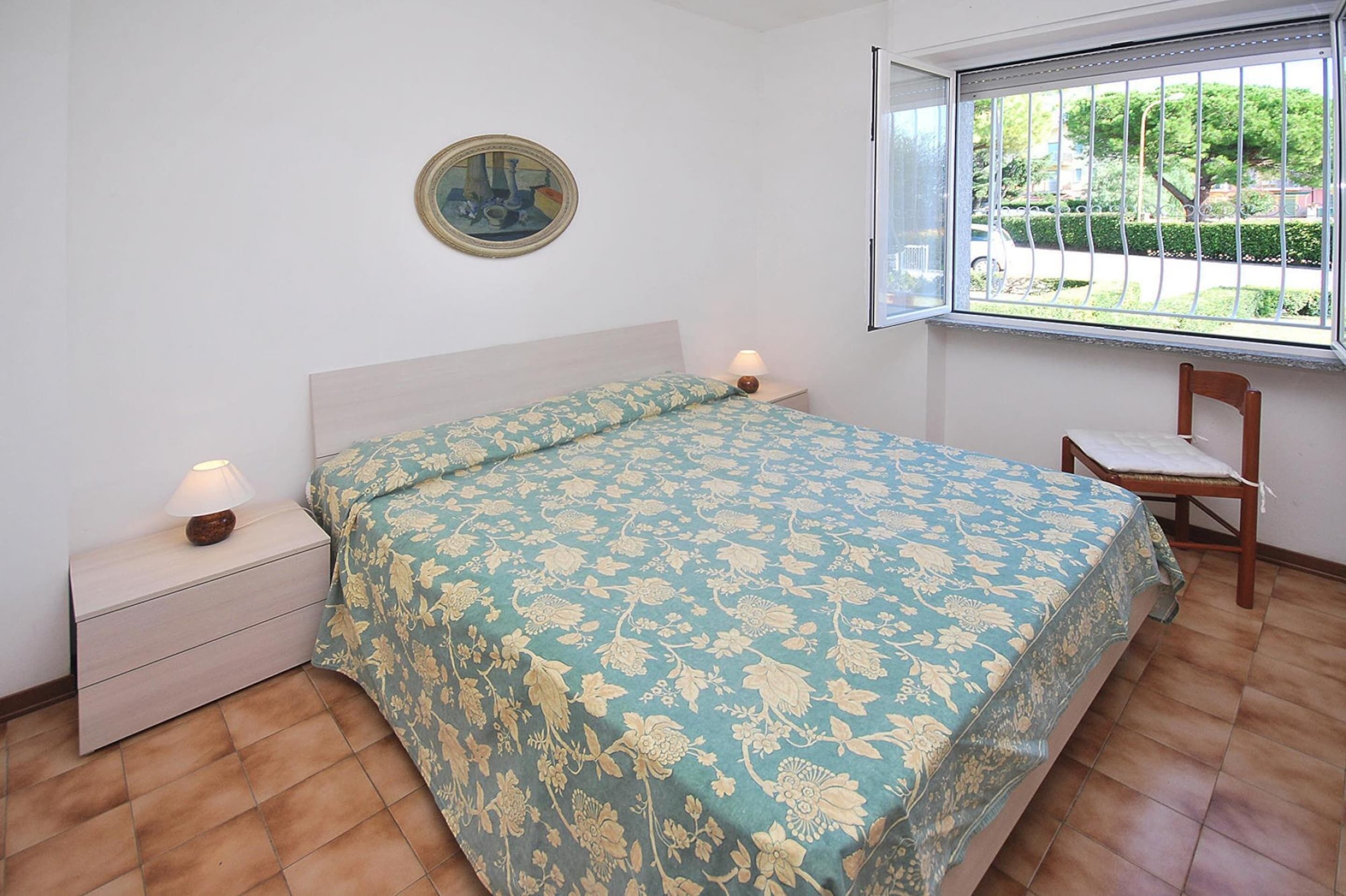 A bedroom with a bed, a nightstand, a chair, a window with a view of the outside, and a painting on the wall.