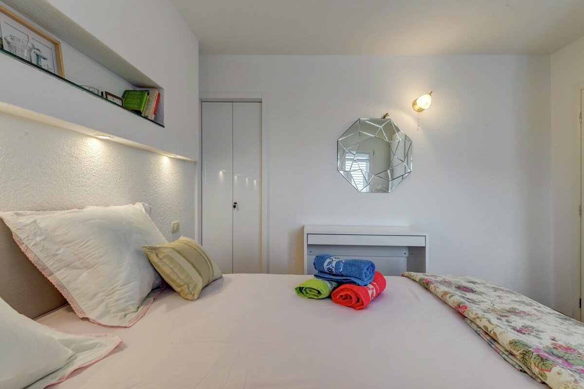 A bedroom with a bed, pillows, a bedside table, a mirror, and a shelf with books.
