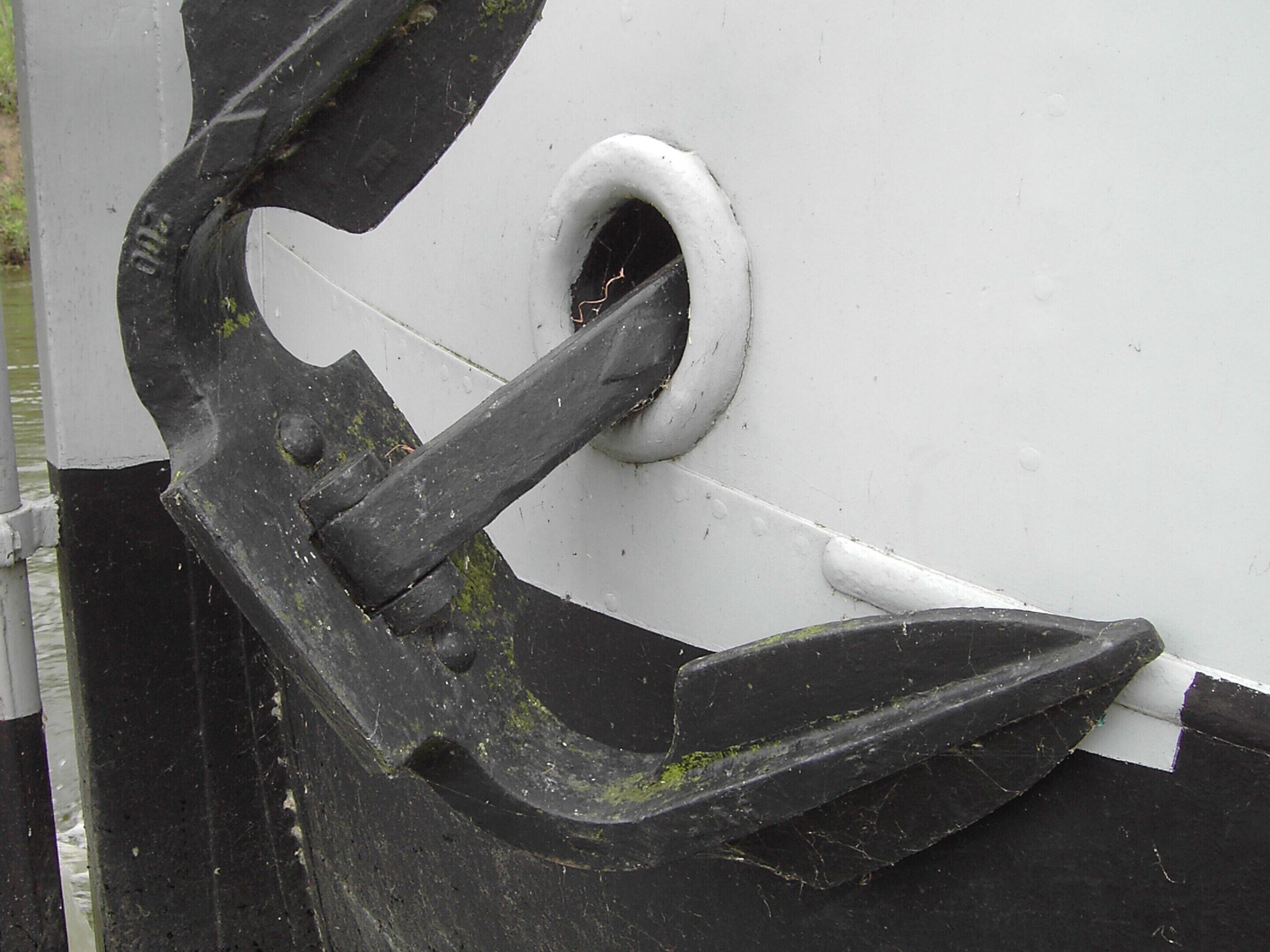 Gros plan d’une ancre métallique encastrée dans une surface blanche et noire, qui fait probablement partie de la structure d’un navire.