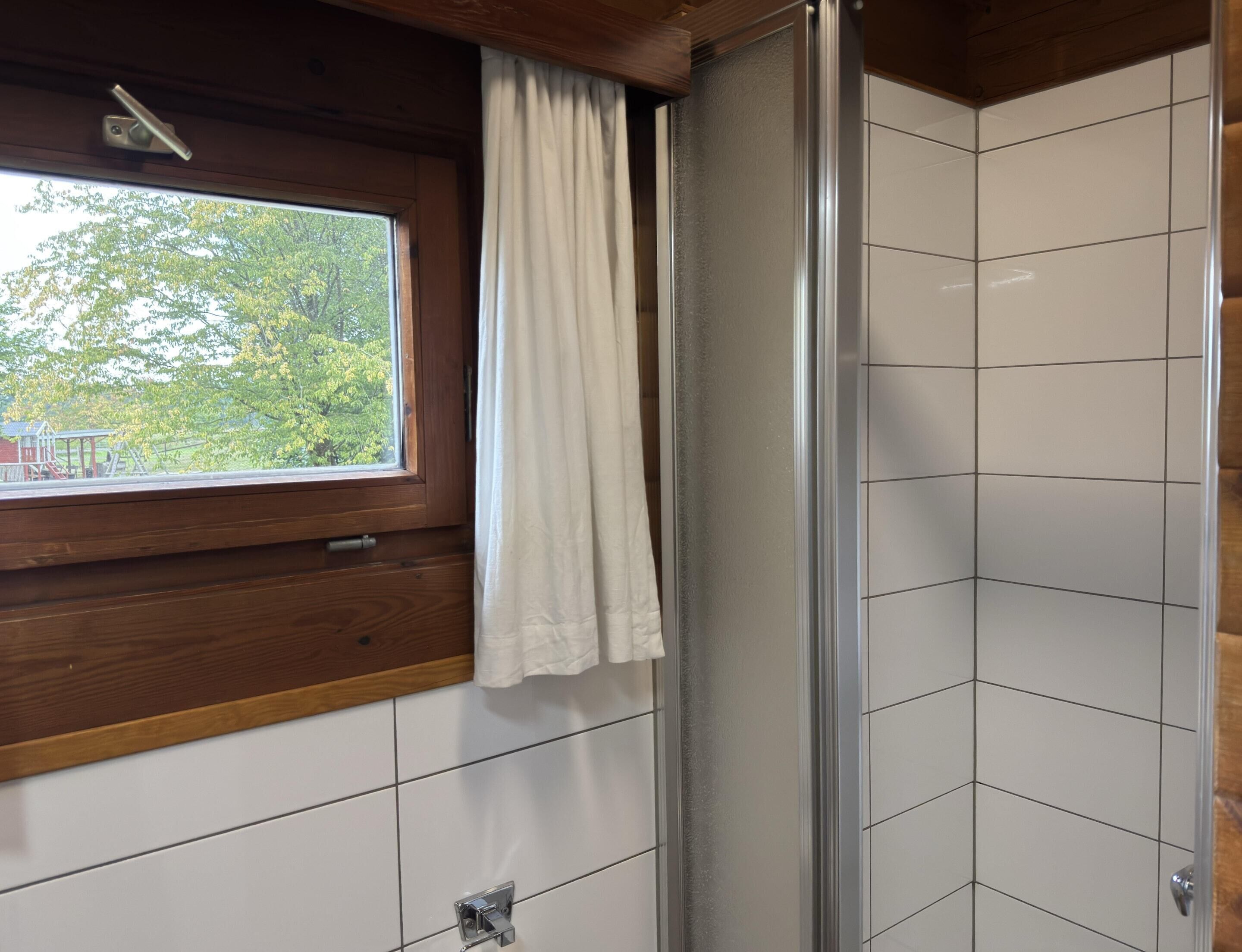 Ein Badezimmer mit weißen Fliesen, einem Fenster mit Vorhang und Blick auf Bäume sowie einen Spielplatz.