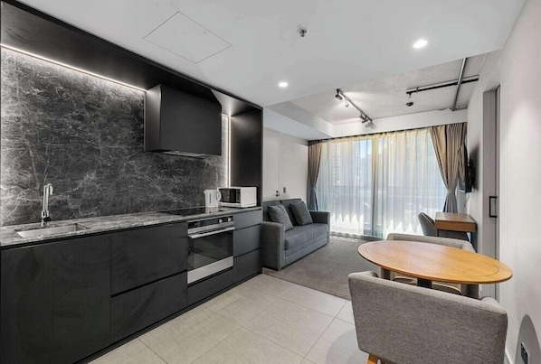 A modern kitchen with a black countertop, built-in oven, and a grey sofa in a living area with a round wooden table.