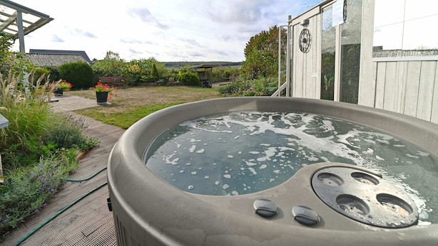A hot tub in a garden with a wooden deck, surrounded by plants and a house in the background.